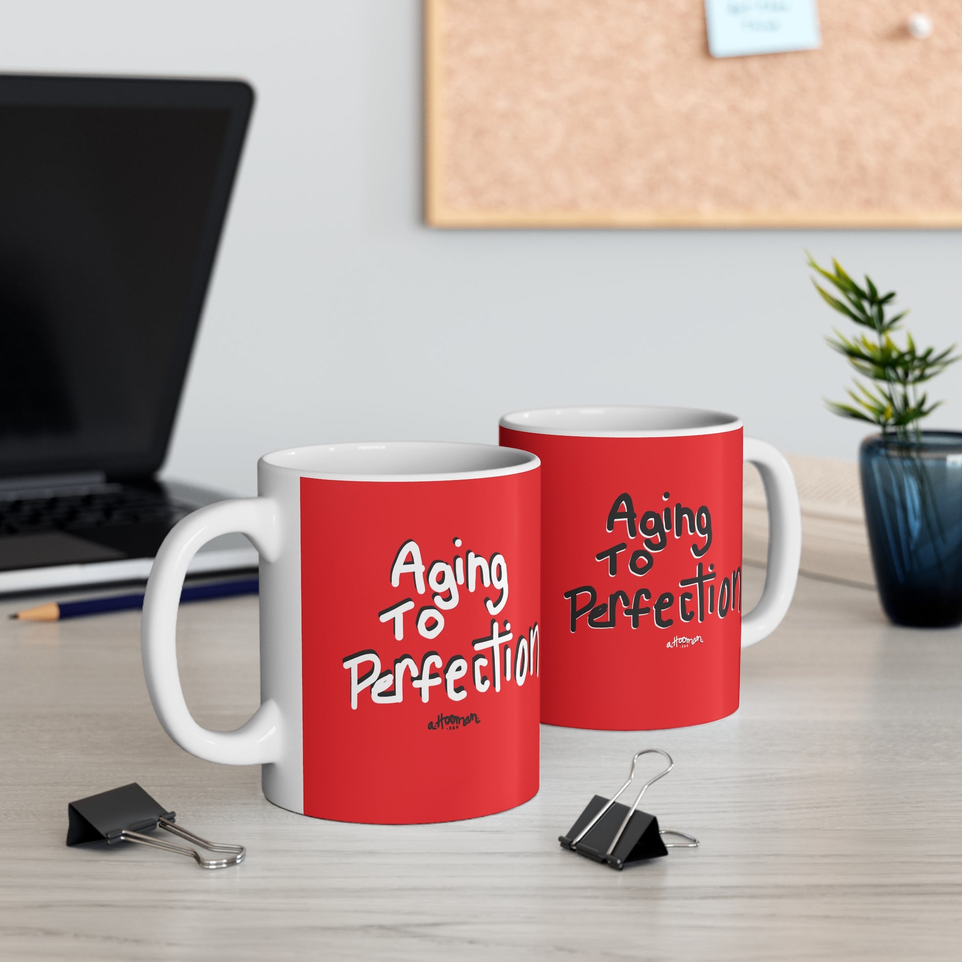 Two red mugs with 'Aging to Perfection' text by a.hooman on a desk with office items.